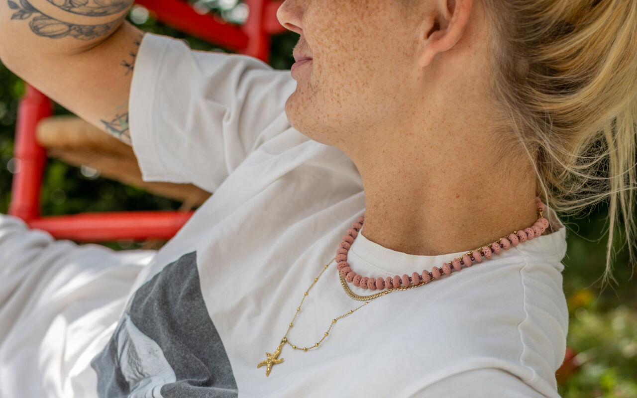 Modelka prezentuje naszyjnik z różowej lawy i granatu w warstwowej stylizacji z pozłacanymi naszyjnikami, tworząc letni, elegancki zestaw.